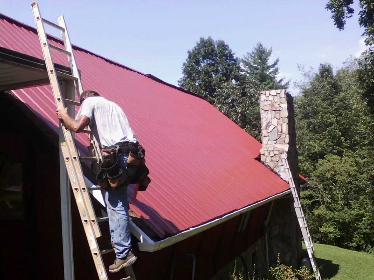 Roofer on a ladder beside a red metal roof and stone chimney — Soffit & Fascia Repair by Roofing Brothers in Chestertown